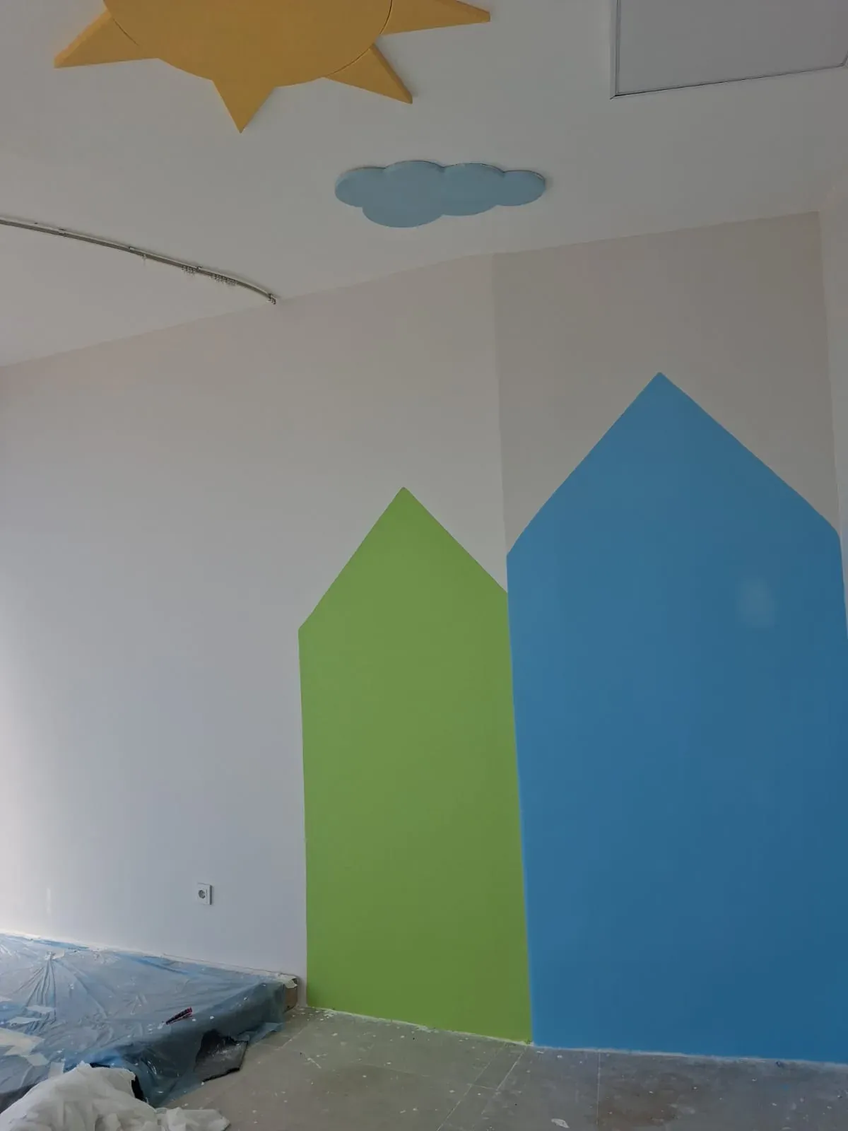 Çocuk odasında ev şeklinde yeşil ve mavi dekoratif duvar boyası, tavanda güneş ve bulut figürleri